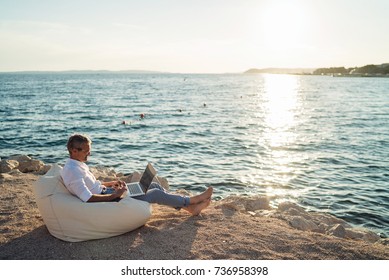 Senior man working on his laptop lying on deck chair on the beach during sunset - Powered by Shutterstock