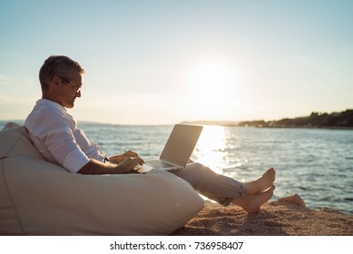 Senior man working on his laptop lying on deck chair on the beach during sunset