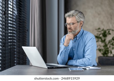 Senior man working on laptop in modern office environment, looking thoughtful. Natural light through window blinds creates a professional and focused atmosphere. - Powered by Shutterstock