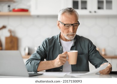 Senior Man Working With Papers And Laptop In Kitchen And Drinking Coffee, Handsome Elderly Businessman Wearing Eyeglasses Reading Documents, Enjoying Remote Work And Business On Retirement - Powered by Shutterstock