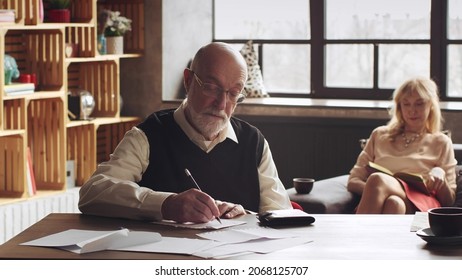 Senior Man Writing A Letter Sitting At Desk In Living Room With Aged Wife Reading Book On Sofa In Background.