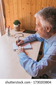 Senior Man Writing In A Notebook At The Wooden Table In His Office, In A Relaxing Environment. Grandfather Writing A Letter To His Grandchildren Or Children During The Quarantine At Home.