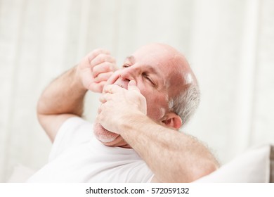 senior man yawning sat on his couch in his living room in an very light coloured home, he's about to go to sleep - Powered by Shutterstock