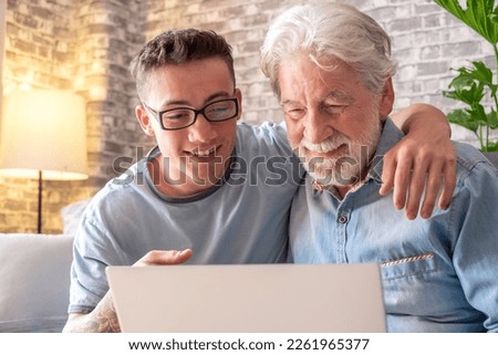 Senior man and young boy smiling and looking together at laptop at home. Happy nephew teaching and showing new computer technology to his old grandfather. Old man learn to use computer.