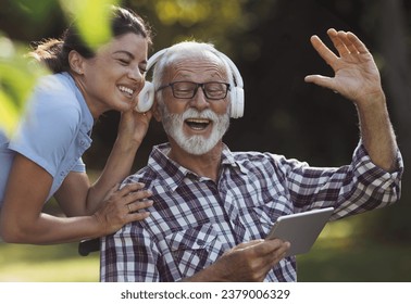 Senior man and young woman nurse listening music from tablet in park on bench and singing  - Powered by Shutterstock