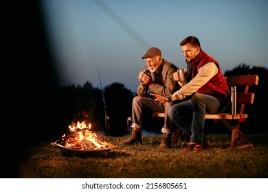 Senior mand and his adult son having a drink while relaxing by bonfire at dusk during their camping weekend.  Copy space. - Powered by Shutterstock