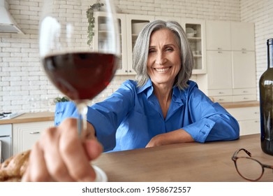 Senior Mature 60s Aged Older Woman Sitting At Kitchen Home At Table With Computer. Having Virtual Wine Party With Friends Family On Remote Video Call Meeting Clinking Glass To Camera, Web Cam View.