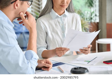 Senior mature Asian business woman boss reading report of young intern trainee in modern office. Teacher checking student's academic work. Middle aged hr director on interview with new coworker.