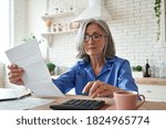Senior mature business woman holding paper bill using calculator, old lady managing account finance, calculating money budget tax, planning banking loan debt pension payment sit at home kitchen table.