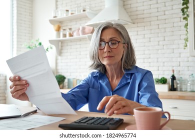 Senior mature business woman holding paper bill using calculator, old lady managing account finance, calculating money budget tax, planning banking loan debt pension payment sit at home kitchen table. - Shutterstock ID 1824965774