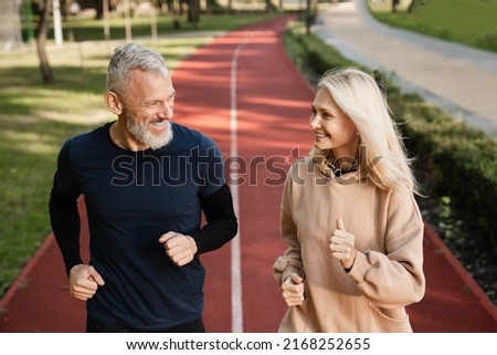 Senior mature couple running together in the park stadium looking at each other while jogging slimming exercises. Training workout