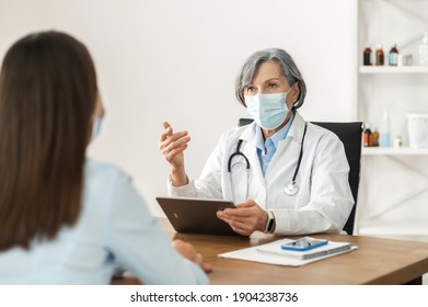 Senior mature female doctor in a lab coat and a face mask sitting at the desk,holding a pad,looking at the young lady patient, explaining medical results, digitized medical records in pandemic concept - Powered by Shutterstock