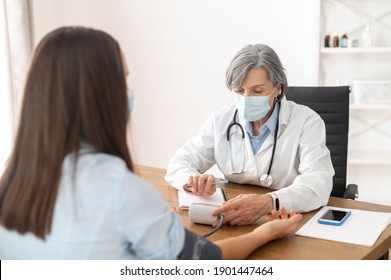 Senior Mature Female Doctor Wearing A Lab Coat And A Face Mask, Using A Blood Pressure Machine Or Sphygmomanometer With Stethoscope To Check A Sick Patient In The Hospital During A Pandemic