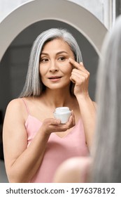 Senior Mature Happy Middle Aged Asian Woman, Older Grey Haired Lady Looking At Mirror Applying Whitening Antiaging Anti Wrinkle Perfect Skin Care Cream To Eye Contour. Headshot.