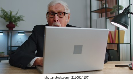 Senior Mature Older Business Man Hiding Behind Laptop Computer, Making Funny Face, Fooling Around, Putting Her Tongue Out Remote Working Disrespecting Someone At Home Office. One Elderly Grandfather