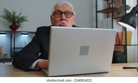 Senior Mature Older Business Man Hiding Behind Laptop Computer, Making Funny Face, Fooling Around, Putting Her Tongue Out Remote Working Disrespecting Someone At Home Office. Elderly Grandfather