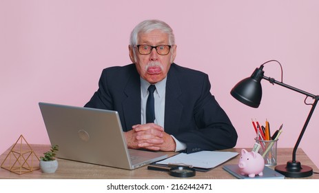 Senior Mature Older Business Man Working On Laptop, Making Funny Face, Fooling Around, Putting His Tongue Out Remote Working Disrespecting Someone At Office. Elderly Grandfather On Pink Background