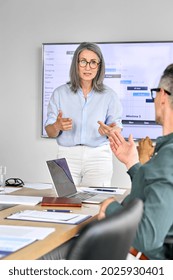 Senior Mature Older Female Executive Ceo Presenter In Glasses Discussing Presentation Of Corporation Financial Global Goals On Big Screen With Executives At Boardroom Modern Office. Vertical.