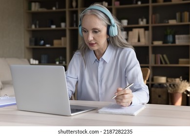 Senior Mature Student Woman In Big Headphones Watching Learning Webinar, Online Class, Lesson On Laptop Computer, Making Video Call, Writing Notes, Getting Knowledge, New Profession At Home