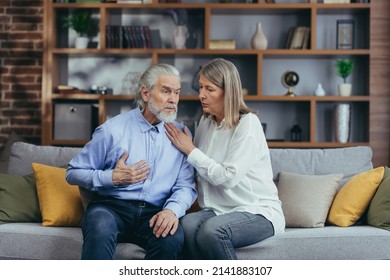 Senior Mature Woman Helping Her Husband With Chest Pain. Old Man Feeling Bad Pain Disease. Wife Supporting Husband - Elderly Couple Suffering From Symptoms Heart Attack Ache. Sitting At Home Indoors