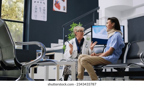 Senior medic checking patient with neck injury while asian assistant writing medical expertise on clipboard during checkup visit in hospital lobby. Injured man wearing cervical collar in waiting area - Powered by Shutterstock