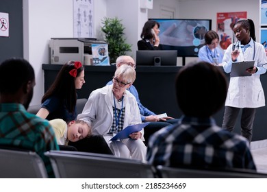 Senior Medical Doctor With Stethoscope Completing Form For Woman And Ill Daughter For Routine Checkup Appointment In Private Practice Clinic. Diverse People Waiting In Modern Hospital Reception.