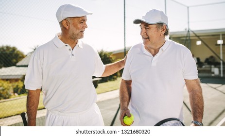 Senior men walking on a tennis court during a game of tennis. Smiling senior men in tennis wear talking to each other holding tennis rackets and ball. - Powered by Shutterstock