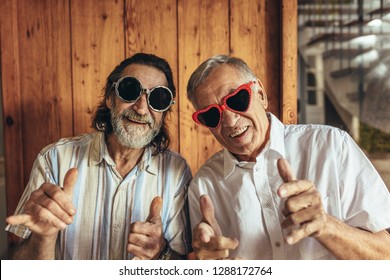 Senior Men Wearing Funny Sunglasses Looking At Camera. Happy Elderly Friends With Crazy Eyewear With Funky Gestures.