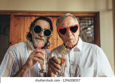 Senior men wearing funny sunglasses drinking juice with straw. Elderly friends with crazy eyewear having juice indoors.