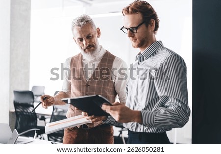 Senior mentor explaining project to young businessman with tablet during office meeting, brainstorming creative business ideas in modern coworking space