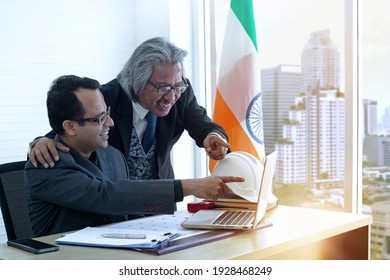 Senior and middle age Indian businessman using computer in the office and happy with good news result of work, modern city buildings can be seen from the windows
