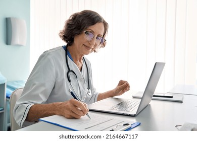 Senior middle aged female medical worker in modern clinic wearing white doctor's coat having videocall using laptop writing health personal data, consulting remotely. Telemedicine healthcare concept. - Powered by Shutterstock