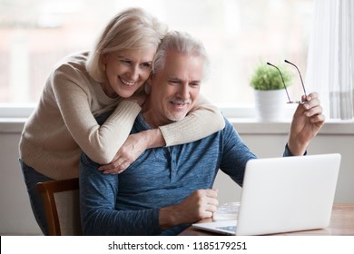 Senior middle aged happy couple embracing using laptop together, smiling elderly family reading news, shopping online at home, older people and computer or good vision after laser correction concept