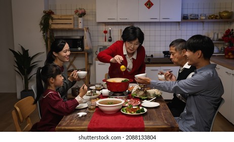 Senior Mistress Of The Family Grandmother Standing Up And Offering Food To Each Member With Chopsticks At Dinner Table During Chinese New Year's Eve Reunion Meal At Home