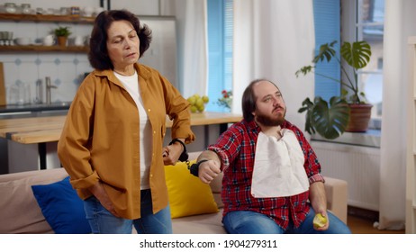 Senior Mother And Adult Son Hands In Handcuffs At Home. Portrait Of Young Overweight Man Sitting On Couch And Eating Apple Handcuffed With Elderly Mother