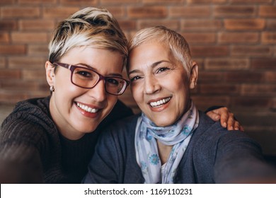 Senior Mother Sitting In Cafe Bar Or Restaurant With Her Middle Age Daughter, They Are Smiling And Taking Selfie Photo.