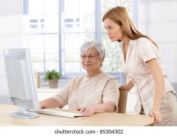 Senior Mother And Young Daughter Using Computer At Home, Looking At Screen.?