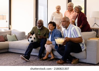 Senior multiracial friends with popcorn and beer watching soccer match in nursing home. Ball, sport, curiosity, alcohol, unaltered, togetherness, support, assisted living and retirement concept.