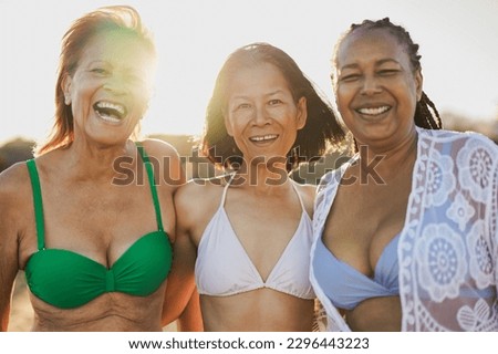 Senior multiracial women having fun hugging each other on the beach and smiling on camera during sunrise - Joyful elderly and travel vacation concept