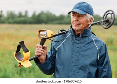 Senior numismatist wearing cap and jacket holding metal detector over his shoulder and looking away, treasure hunter searching historical artifacts.