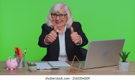 Senior old business woman employee using laptop computer notebook, raises thumbs up agrees with something or gives positive reply recommends advertisement likes good sits at green chroma key office
