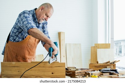 Senior Old Caucasian Man Wearing Check Shirt, Apron, Making DIY Wooden Furniture, Using Equipment To Drill Woods With Happiness While Standing Alone At Indoor Home. Retirement And Hobby Concept