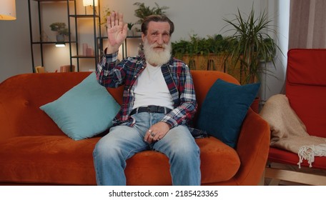 Senior Old Grandfather Man Smiling Friendly At Camera And Waving Hands Gesturing Hello, Hi, Greeting Or Goodbye, Welcoming With Hospitable Expression. Elderly Pensioner Indoor At Home In Room On Couch