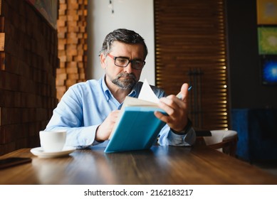 Senior Old Man Reading A Book In A Coffee Shop, Enjoying His Literary Hobby.