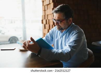 Senior Old Man Reading A Book In A Coffee Shop, Enjoying His Literary Hobby.