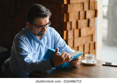 Senior Old Man Reading A Book In A Coffee Shop, Enjoying His Literary Hobby.