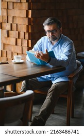 Senior Old Man Reading A Book In A Coffee Shop, Enjoying His Literary Hobby.