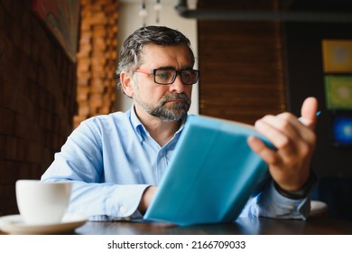 Senior Old Man Reading A Book In A Coffee Shop, Enjoying His Literary Hobby.