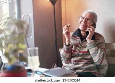 Senior Old Woman Enjoying A Fun Conversation Over Her Cell Phone, Eating A Chocolate Cookie In A Coffee Shop