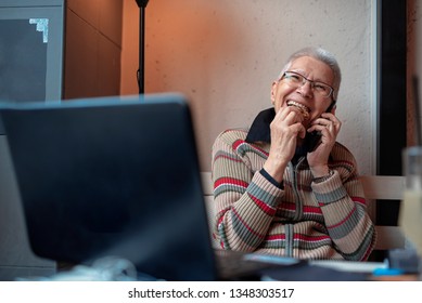 Senior Old Woman Enjoying A Fun Conversation Over Her Cell Phone, Eating A Chocolate Cookie In A Coffee Shop